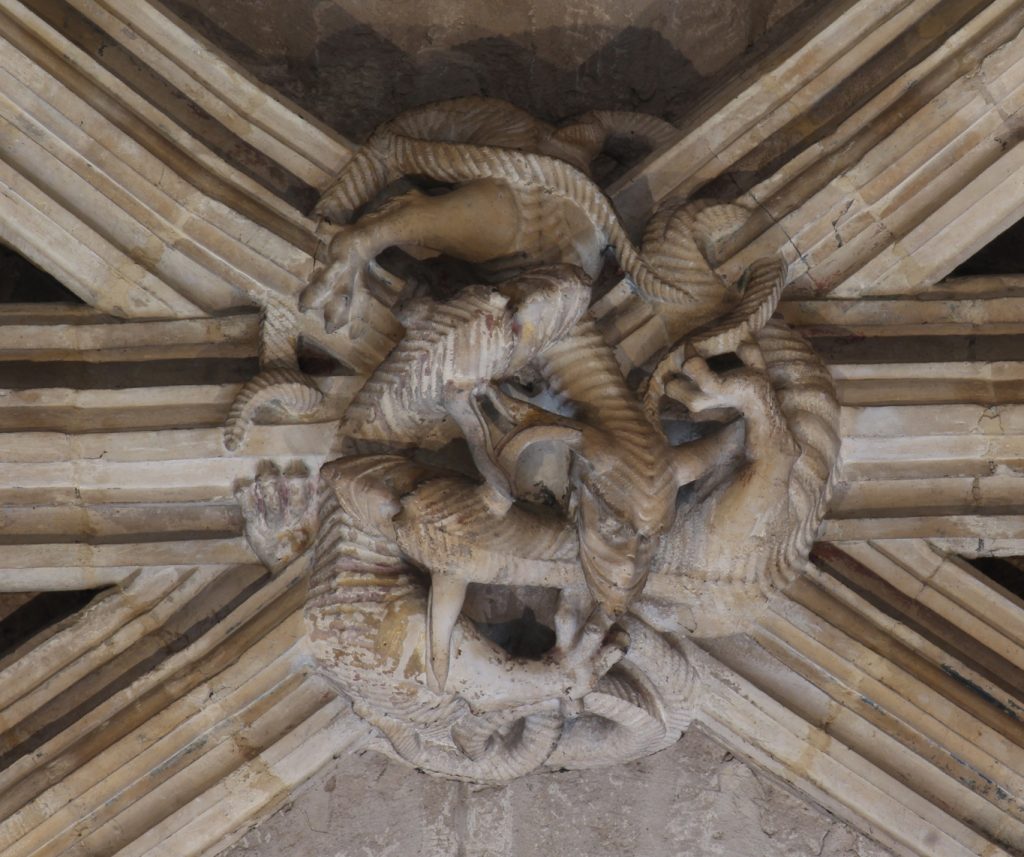 Image of boss in the Angel Choir aisle at Lincoln Cathedral, showing a circular arrangement of three intertwining draconine creatures biting each other's necks and weacing their limbs and tails through the mouldings of the adjoining ribs